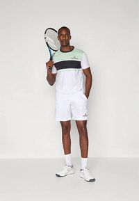 Άνδρας ντυμένος με λευκή και μέντα πράσινη στολή τένις που κρατάει μια ρακέτα τένις πάνω στον ώμο του, στέκεται μπροστά σε ένα απλό φωτεινό φόντο.