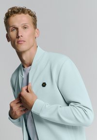 Lichtblauwe bomberjack met een volledige ritssluiting, ribbelmanchetten en een zwart hexagonaal logo op de borst. Gladde stof met een casual ontwerp.