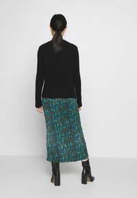 Svart stickad tröja över en mönstrad maxiklänning med gröna och blå nyanser, kompletterad med svarta klackade ankelskålar.