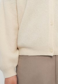 Cardigã bege tricotado com textura canelada, apresentando um decote em V e fechos de botão em creme. Combinado com tecido castanho-claro, exibindo detalhes nas costuras laterais.