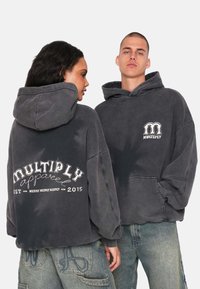 Sweatshirts gris surdimensionnés avec poches avant. Le dos présente "MULTIPLY apparel" en texte blanc. Poignets et ourlet côtelés, texture douce, style décontracté.