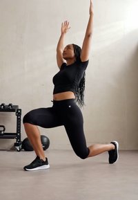 Conjunto deportivo negro, que incluye una parte superior ajustada y leggins hasta la rodilla, usado durante un ejercicio de zancada. Tejido texturizado, combinado con zapatillas negras.