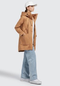 Braune, leichte Jacke mit Kapuze, Reißverschluss und zwei Fronttaschen. Kombiniert mit hellblauen, weit geschnittenen Jeans und weißen Turnschuhen.