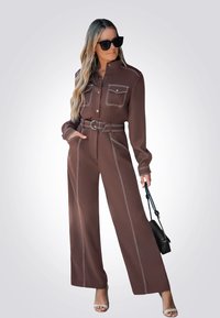 Conjunto marrón de dos piezas con mangas largas, dos bolsillos en el pecho, costuras contrastantes en blanco, pantalones de pierna ancha, cintura ceñida con cinturón y bolso oscuro.