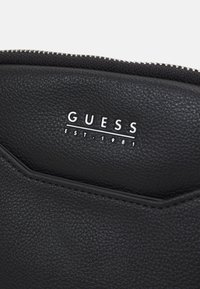 Portafoglio in pelle nera con superficie testurizzata, chiusura a zip e logo "GUESS" in rilievo. Presenta un design elegante e accenti minimalisti.