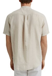 Homme portant une chemise en lin beige à manches courtes et un pantalon blanc, debout de dos avec les bras détendus le long du corps.