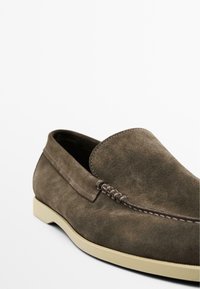 Chaussure en daim marron à enfiler avec un bout arrondi, des coutures contrastantes et une semelle en caoutchouc de couleur claire. Design minimaliste sans matériel visible.