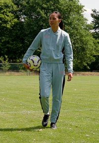 Trening de culoare albastru deschis, cu dungi negre pe laterale, pantofi negri și o minge de fotbal. Teren de iarbă și copaci în fundal.