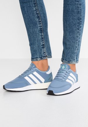 Osoba nosząca niebieskie buty sportowe Adidas z białymi paskami i niebieskie dżinsy, stojąca na białej podłodze na jednolitym tle.
