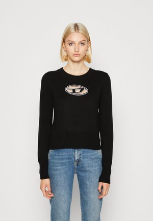 Maglione nero a maglia con scollatura rotonda, polsini e fondo a coste, dotato di una patch con logo beige al centro. Indossato con jeans blu.