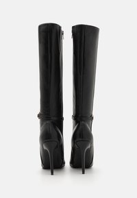 Bottes en cuir noires hautes jusqu'aux genoux avec talons aiguilles, lanières à la cheville et fermetures éclair à l'arrière sur fond blanc neutre.