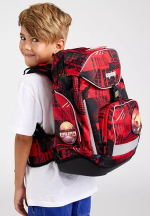 Junge trägt einen großen rot-schwarzen Schulrucksack mit Muster, reflektierenden Elementen und Stadtansicht-Abzeichen an den Seiten.