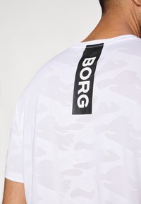 Hvid T-shirt med et diskret camouflage mønster, korte ærmer og et vertikalt sort label med teksten "BORG" på ryggen.