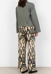 Femme portant une veste grise et un pantalon large avec un motif abstrait noir, vert et beige, debout sur un sol en béton.