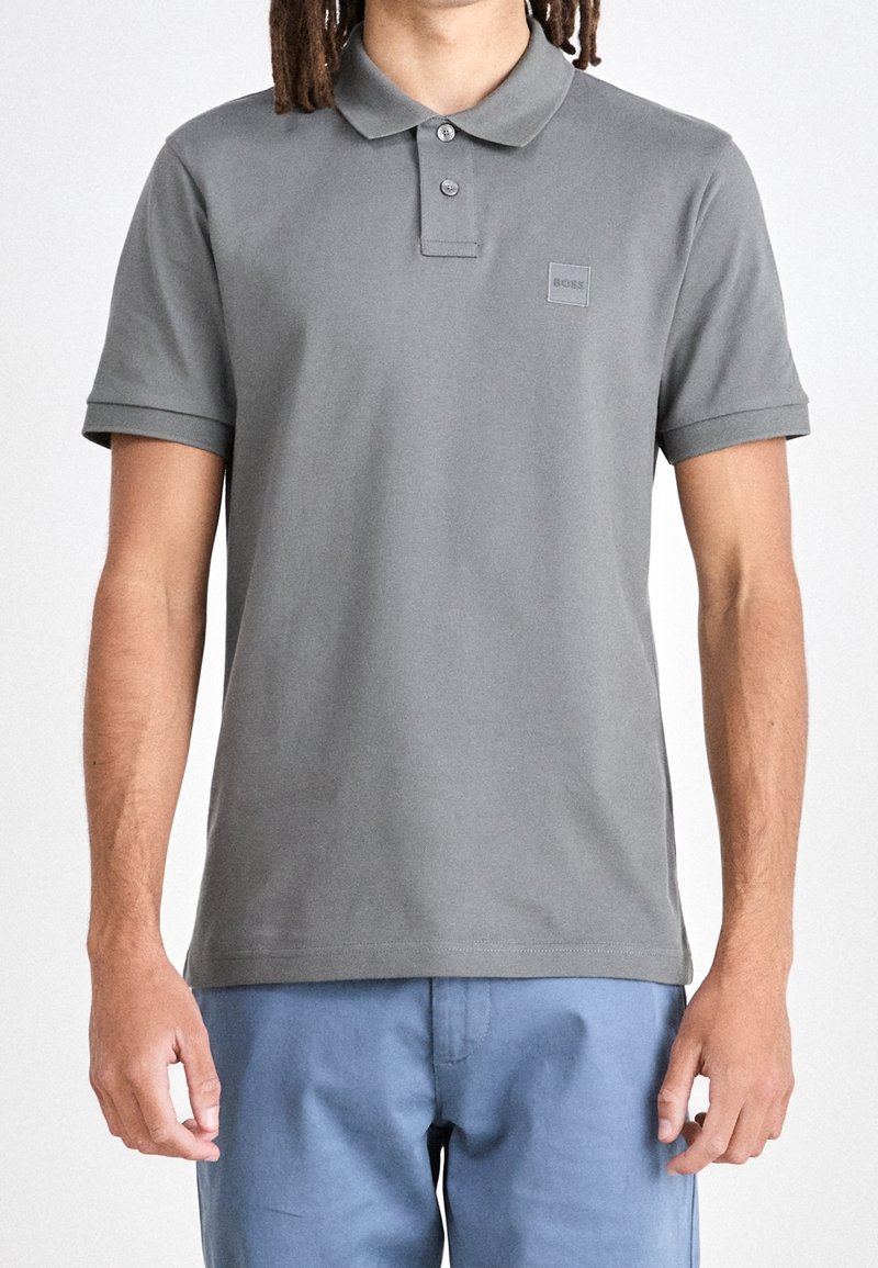 Homme portant un polo gris uni à manches courtes avec deux boutons et un pantalon bleu, debout devant un fond uni.