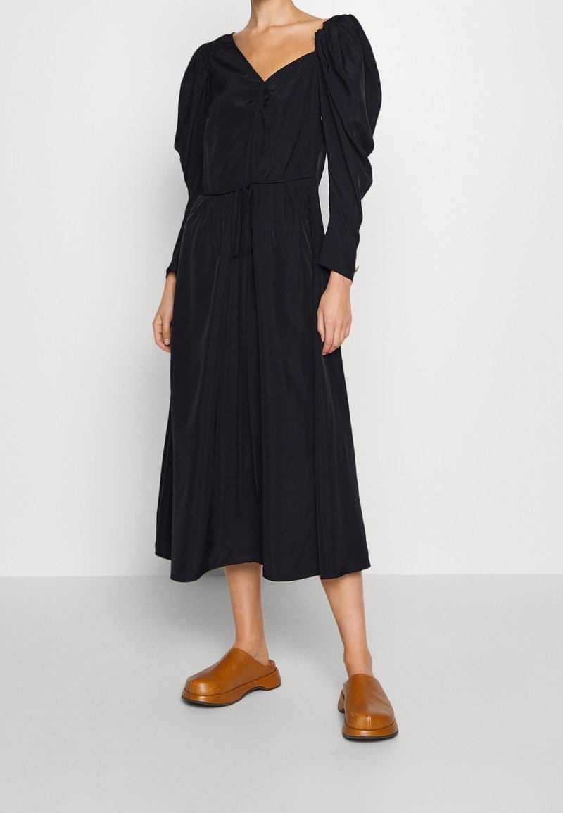 Svart midi-klänning med puffade långa ärmar, V-ringning, draperad midja och knyt detaljer. Kombineras med bruna träträsko. Slätt tyg.