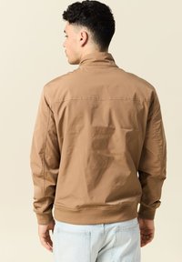 Chaqueta bomber marrón, textura suave, puños y dobladillo acanalados, material ligero, diseño sencillo sin patrones ni detalles de hardware visibles.