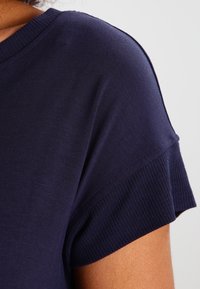 Granatowa koszula z krótkim rękawem z ribbowanym dekoltem i wykończeniem rękawów. Gładka tekstura z luźnym krojem. Widoczny detal szwu na ramieniu.