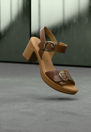 Sandale marron à talon épais, avec deux sangles ajustables et de grandes boucles métalliques. Tige en cuir lisse, semelle plateforme texturée.