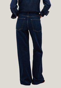 Persona vestida con pantalones de mezclilla de pierna ancha en azul oscuro y camisa de mezclilla a juego, de pie con las manos en las caderas contra un fondo claro.