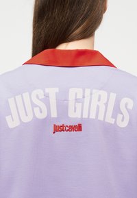 Γυναίκα με μακριά καστανά μαλλιά, ντυμένη με πουκάμισο λεβάντας με κόκκινο γιακά, με λευκό κείμενο "JUST GIRLS" και κόκκινο "Just Cavalli" στην πλάτη.