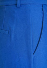 Modré kalhoty mají hladkou, přiléhavou látku s elegantním designem, včetně boční kapsy a strukturovaného pásku.