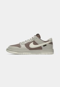 Bež i smeđe Nike Dunk Low tenisice izrađene od kože i tekstilnih materijala. Imaju zaobljeni vrh, vezice i kontrastni logotip sa strane.