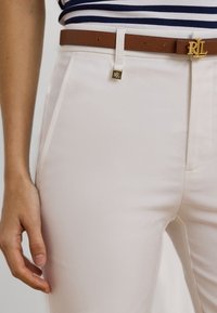 Ljusbeiga byxor med en slät textur, utrustade med ett brunt läderbälte med ett guldspänne. Vertikala fickor är synliga fram.