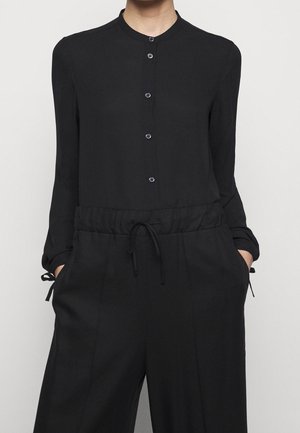 Personne portant une chemise noire à manches longues boutonnée et un pantalon noir avec taille à cordon, mains dans les poches, sur un fond uni.