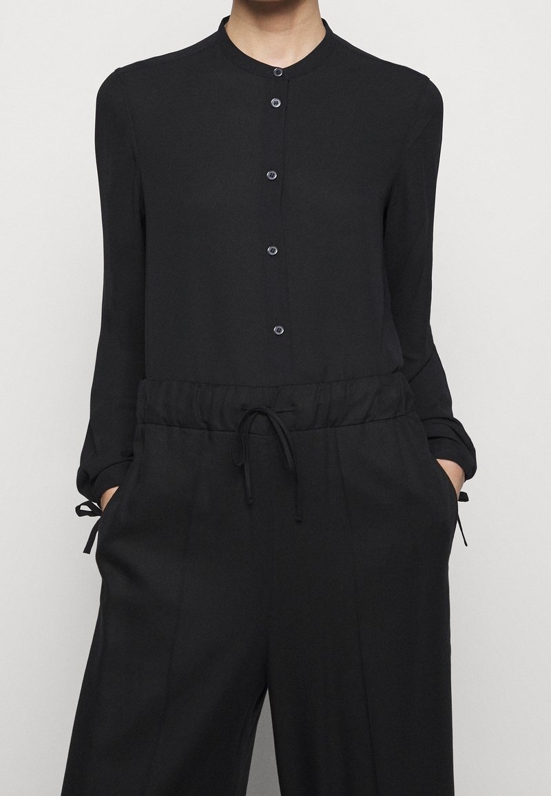 Personne portant une chemise noire à manches longues boutonnée et un pantalon noir avec taille à cordon, mains dans les poches, sur un fond uni.