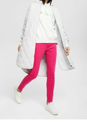 Vrouw draagt een witte gewatteerde jas, een witte trui met meerkleurige tekst, felroze broek en witte sneakers loopt vooruit.