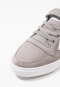 Primer plano de la parte delantera de una zapatilla deportiva gris con parte superior de ante y tejido, cordones a juego, ojales de metal y una suela de goma blanca.