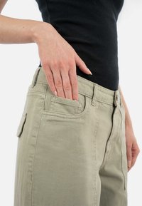 Světle olivově zelené kalhoty s hladkým povrchem, standardními kapsami a přiléhavým střihem. Ruka je umístěna uvnitř přední kapsy.