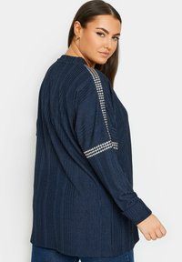 Marinblå långärmad topp med ribbad textur och silverfärgade dekorativa detaljer längs ärmarna. Löst snitt med rund halsringning och sprund på sidan.