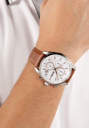 Montre-bracelet avec cadran blanc, trois sous-cadrans, affichage de la date, boîtier argenté et bracelet en cuir marron perforé porté au poignet gauche d'une personne.