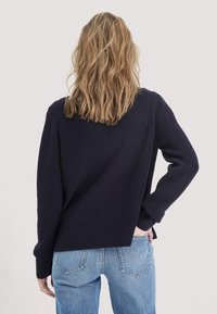 Vrouw met golvend blond haar, gekleed in een donkerblauwe trui en lichtblauwe jeans, met één hand op haar heup en met haar rug naar de kijker gekeerd.