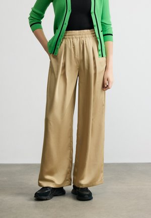 Persona valkā bēša krāsas bikses ar elastīgu jostu, melnu augšu, zaļu džemperi ar pogām un melnus sporta apavus, stāv uz pelēkas grīdas.