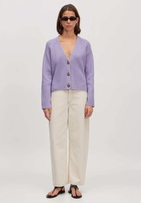 Paarse gebreide cardigan met een V-hals en vijf bruine knopen, gecombineerd met roomkleurige wijde broeken en zwarte slippers.