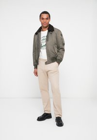 Oliven grøn bomberjakke med pelskrave, hvid grafisk t-shirt, beige bukser og sorte loafers. Model står foran en hvid baggrund.