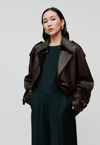 Braune Lederjacke mit großem Kragen, Reißverschlüssen und Knopfdetails, über einem dunkelgrünen Kleidungsstück getragen. Modell mit langen Haaren und goldenen Ohrringen.