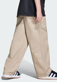 Beżowe spodnie o szerokich nogawkach z trzema pionowymi ciemnymi paskami. Materiał wydaje się wytrzymały, zestawiony z czarnymi sportowymi butami z białymi akcentami.