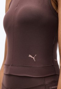 Κοντινό πλάνο ενός ατόμου που φοράει καφέ αθλητική μπλούζα τύπου τοπ και κολάν Puma με ορατή ραφή και λογότυπο σε λευκό φόντο.