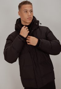 Svart polstret jakke med høy krage og justerbar hette, med glidelås og trykknapper foran, glatt tekstur og quiltet design.