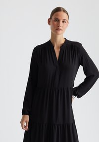 Schwarzes Kleid mit langen Ärmeln aus weichem Stoff, mit hohem Ausschnitt, gerafften Details an den Schultern und einem gestuften Rockdesign.