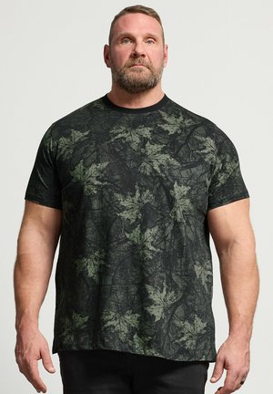 Brodaty muskularny mężczyzna stoi nieruchomo, nosząc ciemnozieloną koszulkę z krótkim rękawem w liściasty wzór kamuflażu na jednolitym tle.