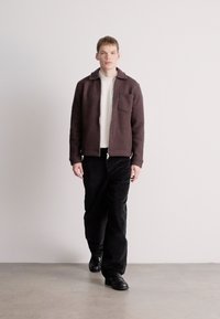 Καφέ υφασμάτινο σακάκι με φερμουάρ μπροστά και τσέπη, φορεμένο πάνω από ένα λευκό πλεκτό πουλόβερ, συνδυασμένο με μαύρο παντελόνι κοτλέ και μαύρα παπούτσια.