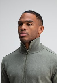 Grijze zip-up sweater met een hoge kraag, geribbelde accenten, een soepele textuur en contrasterende ritsdetails. Close-up van de voorkant van de halslijn.