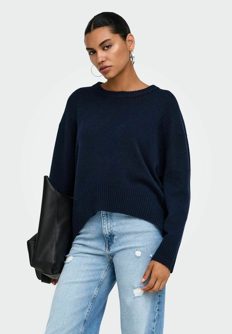 Femme portant un pull bleu marine et un jean clair déchiré tenant un grand sac en cuir noir sur un fond uni.