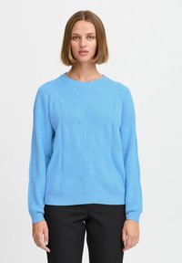 Maglione blu lavorato a maglia con maniche lunghe, scollo tondo, motivi testurizzati e polsini a coste. Indossato sopra pantaloni neri.