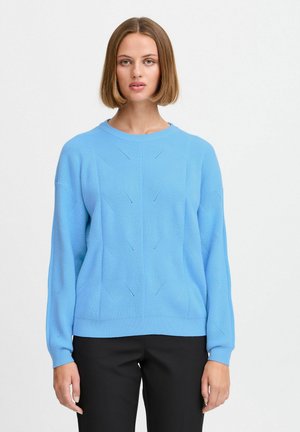 Maglione blu lavorato a maglia con maniche lunghe, scollo tondo, motivi testurizzati e polsini a coste. Indossato sopra pantaloni neri.